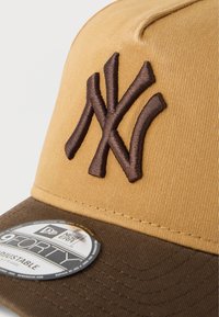 Καπέλο μπεζ με καφέ γείσο, που φέρει χαρακτηριστικό καφέ κεντητό λογότυπο New York Yankees. Ρυθμιζόμενο μέγεθος με κυκλικό ετικέτα στο γείσο.