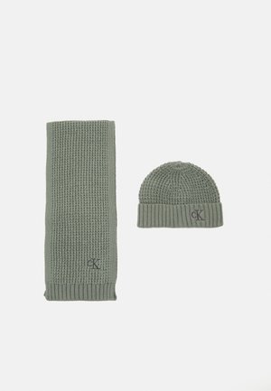 Žalias megztas šalikas ir atitinkantis kepurė. Šalikas turi raštą su gumbeliais; kepurė pasižymi tekstūrinio, prigludusio dizaino. Abu turi logotipo detalę.