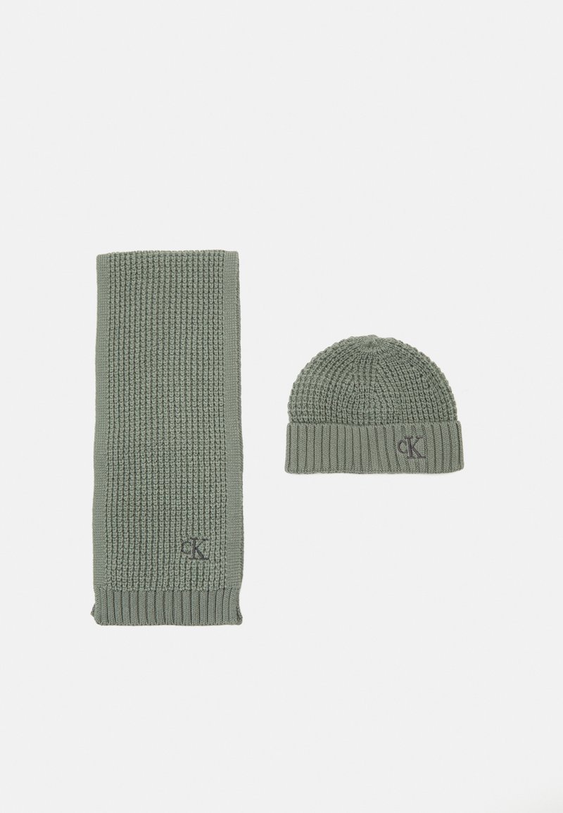 Strikket grønn skjerf og matchende lue. Skjerfet har et ribbestrikket mønster; luen har en strukturert og tilpasset design. Begge har en logodetalj.