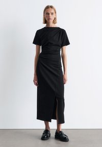 Schwarzes Kleid mit kurzen Ärmeln, taillierter Oberseite und einem seitlichen Schlitz. Glattes Material mit minimalistischer Gestaltung. Kombiniert mit schwarzen Plateauschuhen.