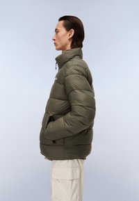 Puffer jacket in olive green, featuring a high collar, quilted texture, zippered front, and side pockets. Simplistic design with minimal branding.