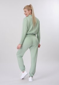 Νεαρή γυναίκα με μακριά ξανθά μαλλιά σε αλογοουρά, φοράει ανοιχτό πράσινο σετ φόρμας και λευκά αθλητικά παπούτσια, στέκεται πλάτη και χαμογελάει.