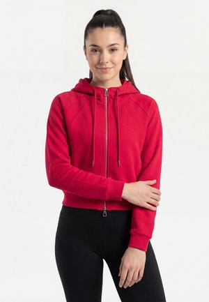 Nuori nainen, jolla on tummat hiukset, pukeutuneena punaiseen vetoketjuhuppariin ja mustiin leggingsseihin, seisoo yhdellä kädellä ristissä vartalonsa edessä.
