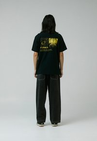 Camiseta de algodón negra con mangas cortas, con estampados gráficos dorados en la espalda. Combinada con pantalones vaqueros de pierna ancha de mezclilla oscura y zapatillas deportivas.