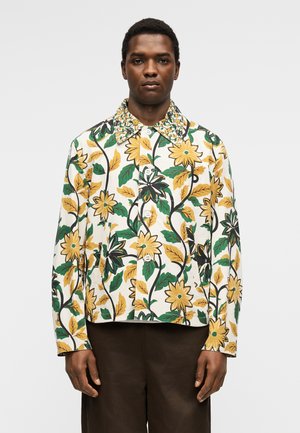 Mand iført en knappet jakke med stor gul og grøn blomsterprint og brune bukser mod en ensfarvet lys baggrund.