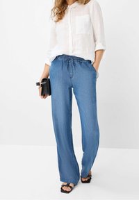 Mujer vestida con una camisa blanca de manga larga y unos jeans azules holgados, sosteniendo un bolso de mano negro y usando sandalias negras de tacón abierto.