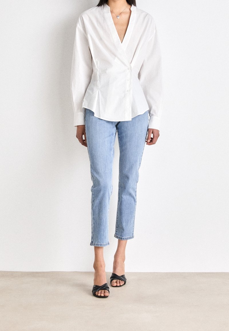 Kvinna som bär en vit omslagsblus, ljusblå cropped jeans, svarta öppna klackskor, stående mot en vit vägg.