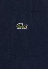 Navy blue fabric featuring a small, embroidered green crocodile logo outlined in white. Smooth texture with a subtle pattern.