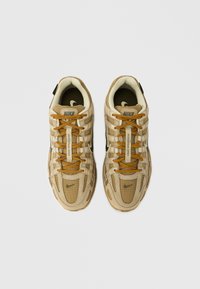 Bézs színű sportcipő hálós és bőr anyagokból. Narancssárga fűzőkkel, fekete díszítésekkel és texturált gumitalppal rendelkezik. Nike márkajelzés található a nyelvén és az oldalán.