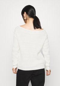 Witte gebreide trui met een brede off-the-shoulder halslijn en geribbelde manchetten. Draag het met zwarte broek, met een gestructureerde, zachte afwerking.