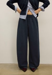 Dunkelblaue, weit geschnittene Hosen mit elastischem Bund, seitlichen Taschen und glatter Textur, kombiniert mit einem hell gestreiften Hemd und dunklen Absätzen.