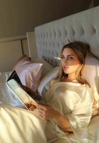 Silkesvitt morgonrock, mjuka beige och rosa kuddar, elegant tuftad sänggavel, tidning i handen som visar modebilder med ett klart, naturligt ljus.