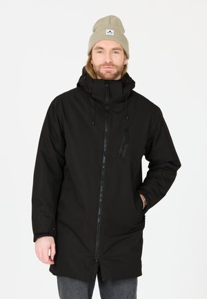 Veste imperméable noire avec un col montant, fermeture éclair à l'avant, poches latérales et capuche avec cordon de serrage. Portée avec un bonnet en tricot beige.