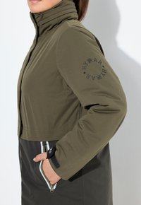 Mujer vestida con una chaqueta oliva y negra con cuello alto, mano en el bolsillo con cremallera y logo circular "HYRAR" en la manga.