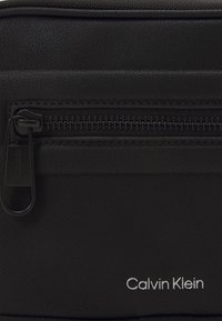Borsa nera in ecopelle con superficie testurizzata. Presenta una tasca con zip e il logo "Calvin Klein" stampato in argento sulla parte anteriore.