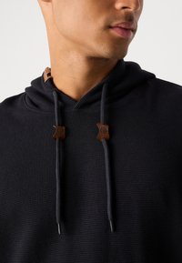 Nahaufnahme einer Person, die einen schwarzen Hoodie mit Kordeln trägt, der braune Ledertabs und geprägte Logos aufweist.