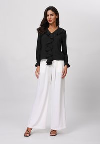 Schwarze Bluse mit gerafftem Vorderteil, V-Ausschnitt und langen Ärmeln; kombiniert mit hochtaillierten, weit geschnittenen weißen Hosen; schwarze Riemchenabsätze.