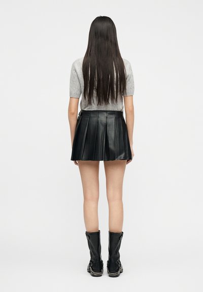 Femme aux cheveux longs et raides portant un haut gris à manches courtes, une jupe plissée en cuir noire et des bottes noires mi-mollet, debout tournée de dos.