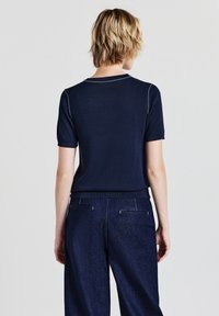 Persona con capelli biondi corti indossa un top a maglia blu navy a maniche corte e pantaloni scuri blu navy abbinati, voltata di spalle su uno sfondo chiaro e uniforme.