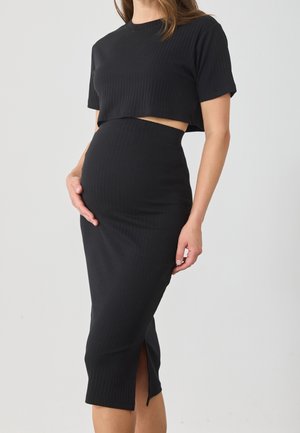 Zwangere vrouw in een aansluitende zwarte geribbelde jurk met een kort mouwcropped top, die met één hand haar buik vasthoudt.