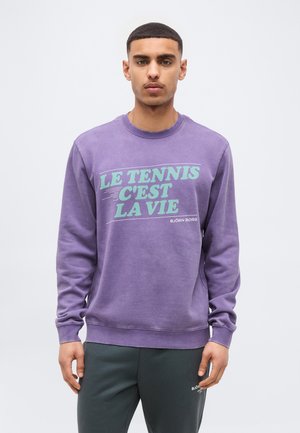 Giovane uomo che indossa una felpa viola con la scritta "LE TENNIS C'EST LA VIE" e pantaloni scuri, in piedi contro uno sfondo chiaro e uniforme.