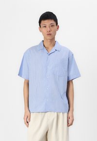 Chemise à manches courtes boutonnée en bleu clair avec des rayures blanches verticales, comprenant une poche poitrine et une coupe décontractée. Textile doux.