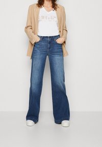 Beige pinstripe bleiseri valkoisen V-pääntie t-paidan päällä, jossa on kohotettu teksti, yhdistettynä leveälahkeisiin haalistuneisiin sinisiin farkkuihin ja valkoisiin lenkkareihin.