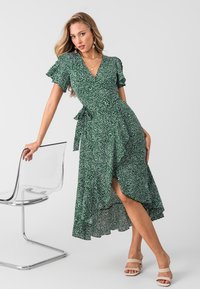 Vestido verde con lunares blancos, manga corta con volantes, dobladillo asimétrico y un lazo en la cintura. Llevado con sandalias beige.