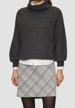 Mujer con jersey de cuello alto texturizado gris oscuro, camisa blanca debajo y falda blanca con patrón de cuadros y medias negras.