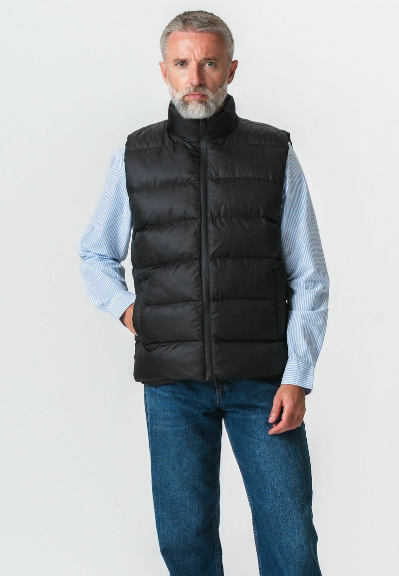 Homme aux cheveux gris et à la barbe, portant un gilet matelassé noir sur une chemise rayée bleue et un jean bleu, debout avec une main dans la poche.