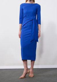 Robe bleue, longueur genou en tissu extensible. Présente des manches trois-quarts et une ceinture à la taille, complétée par des sandales à talons hauts beiges.