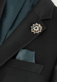Veste de costume noire texturée avec pochette à motif géométrique bleu sarcelle et broche en or comportant une pierre centrale bleue et de petites pierres transparentes.