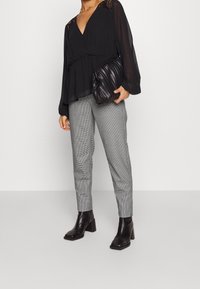 Blouse noire transparente à manches longues, décolleté en V, accents en dentelle ; pantalon gris à carreaux, fuselé ; bottines noires à talons ; sac à main texturé.