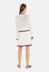Weißes gestricktes Kleid mit einem gerafften Saum und marineblauen Akzenten, ausgestattet mit langen Ärmeln und einem runden Ausschnitt, das eine offene Webstruktur aufweist.