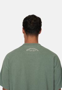 T-shirt in cotone verde con colletto rotondo, maniche corte e branding sul retro in testo bianco, caratterizzato da un taglio oversize e una texture morbida.