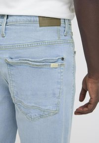 Persona que lleva vaqueros de mezclilla azul claro y una camisa blanca, mostrando el detalle del bolsillo trasero y parte del brazo derecho y la mano.