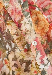 Blouse florale avec un design plissé aux nuances de rose, orange et vert, présentant un tissu léger et un seul bouton blanc.