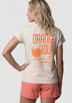 Frau mit welligem blondem Haar trägt ein cremefarbenes T-Shirt mit orangefarbenem Schriftzug und Grafik "ORANGE HOUR keepin' it spritzy", kombiniert mit korallenfarbenen Shorts.