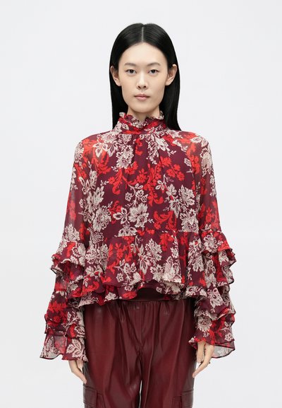 Blouse transparente à motifs floraux en rouge profond et crème, dotée d'un col montant à volants et de manches superposées, confectionnée dans un tissu léger et fluide.