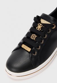 Black sneaker with a textured leather upper, gold-tone hardware on laces and branding, contrasting white sole, and black laces.