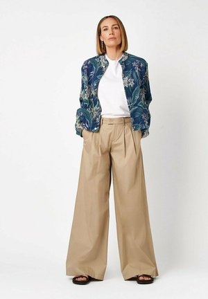 Woman with straight blonde hair wears blue floral jacket, white shirt, wide beige pants, and black sandals, standing against white background.