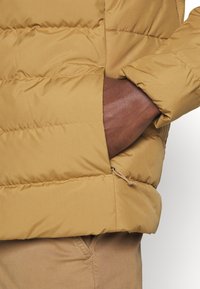 Beige Pufferjacke mit gepolstertem Design und verstellbarem Saum. Die linke Hand befindet sich in einer Seitentasche, die eine glatte Stofftextur zeigt.