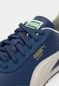 Tamno plave tenisice s kožnim i nubuk teksturama, s crnim vezicama, bijelim prugama i zlatnim logotipom koji glasi "PUMA ARIZONA."