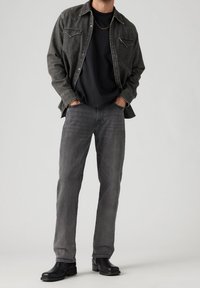 Camicia di denim grigia con due tasche sul petto, indossata sopra una t-shirt nera. Jeans grigi e stivali neri completano l'outfit.