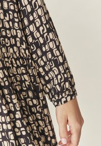 Juoda ilgaa rankovė su smėlio spalvos abstrakčiu raštu. Turi susirinkusią liemenį ir prigludusius rankogalvius. Audinys atrodo minkštas ir tekstūruotas.