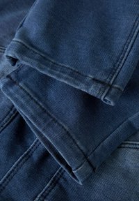 Tejido de denim en tonos azul oscuro, con costuras visibles, puños doblados y ligeras variaciones de textura entre las diferentes capas de tela.