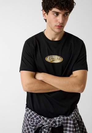 Jeune homme aux cheveux courts et bouclés, debout les bras croisés, portant un t-shirt noir Guess et une chemise à carreaux nouée autour de la taille sur un fond uni.