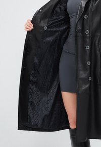 Cappotto in pelle nera con chiusura a bottoni, caratterizzato da un'esterno liscio e una fodera interna nera a motivo con disegni circolari.