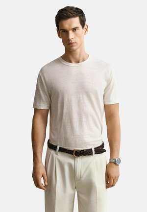 Man met een effen wit T-shirt met korte mouwen, ingestopt in crèmekleurige broek met een gevlochten bruine riem en een zilveren polshorloge.