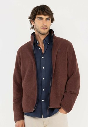 Hombre con una chaqueta de forro polar marrón con cremallera sobre una camisa azul oscuro con botones y pantalones beige claros con una mano en el bolsillo.